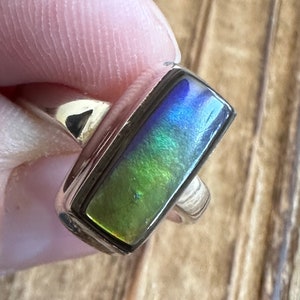 K&ouml;nnte beinhalten: Ein silberner Ring mit einem rechteckigen, schillernden Opal-Edelstein. Der Opal zeigt Farbt&ouml;ne in Gr&uuml;n, Blau und Lila.
