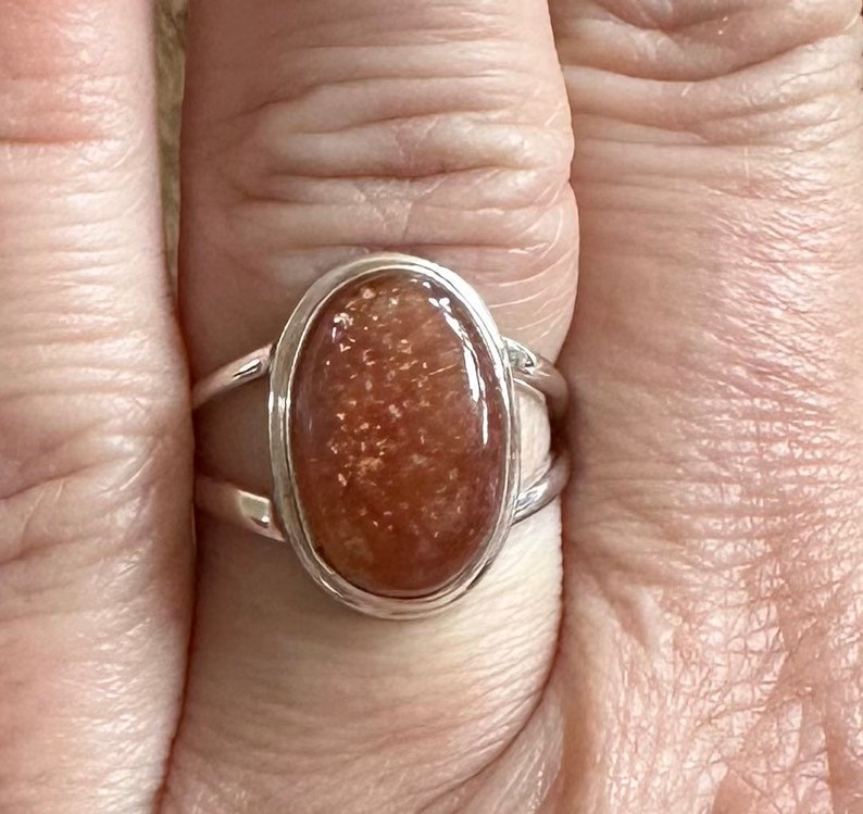 May include: A silver ring with an oval-shaped sunstone. The sunstone is a deep orange color with a shimmery, glittery texture.