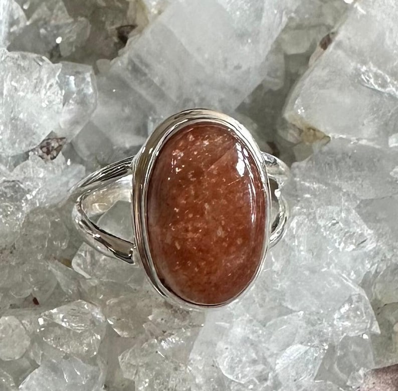 May include: A silver ring with a large oval-shaped sunstone. The sunstone is a reddish-brown color with a shimmery, iridescent finish.