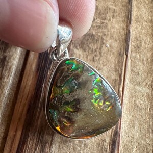 May include: A silver pendant with a dark brown, green, and iridescent opal stone. The stone is set in a silver bezel and has a teardrop shape.