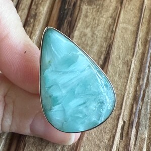 May include: A large, teardrop-shaped ring featuring a light blue gemstone with white marbling. The stone is set in a silver-colored metal band. The ring is displayed against a wooden background.