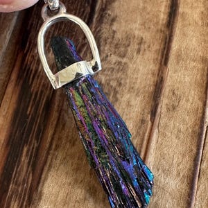 May include: A silver pendant featuring a dark iridescent crystal with a silver bail. The crystal is long and narrow, with a pointed tip. The pendant is perfect for adding a touch of sparkle to any outfit.