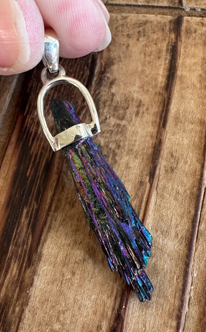 May include: A silver pendant featuring a dark iridescent crystal with a silver bail. The crystal is long and narrow, with a pointed tip. The pendant is perfect for adding a touch of sparkle to any outfit.