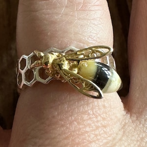 May include: A silver ring with a gold bee design. The bee is sitting on a honeycomb pattern. The ring is on a person's finger.