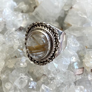 Pu&ograve; includere: Un anello d'argento con una grande pietra ovale trasparente con inclusioni dorate. L'anello ha un bordo intrecciato dettagliato e una fascia ampia e lucida. L'anello &egrave; esposto su un letto di cristalli trasparenti.