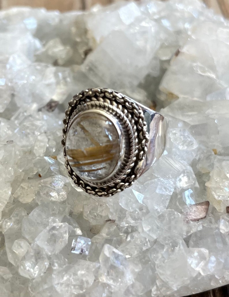 Pu&ograve; includere: Un anello d'argento con una grande pietra ovale trasparente con inclusioni dorate. L'anello ha un bordo intrecciato dettagliato e una fascia ampia e lucida. L'anello &egrave; esposto su un letto di cristalli trasparenti.