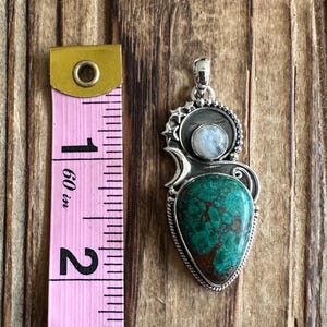 May include: A silver pendant with a green and brown turquoise stone and a moonstone. The pendant is about 2 inches long.
