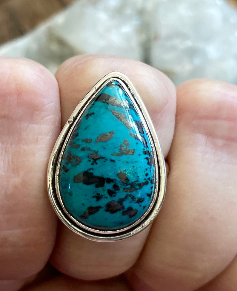 May include: A silver ring with a teardrop-shaped turquoise stone. The stone has a mottled pattern of black and blue.
