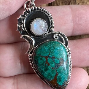 May include: A silver pendant with a green and brown gemstone and a white moonstone. The pendant is intricately designed with a swirl pattern and bead accents.