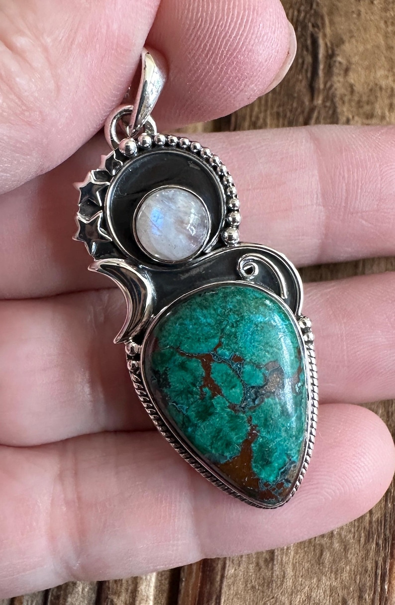 May include: A silver pendant with a green and brown gemstone and a white moonstone. The pendant is intricately designed with a swirl pattern and bead accents.