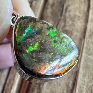 May include: A teardrop-shaped pendant with a silver setting and a dark green, iridescent gemstone. The gemstone has a mottled pattern with flashes of green, orange, and red.