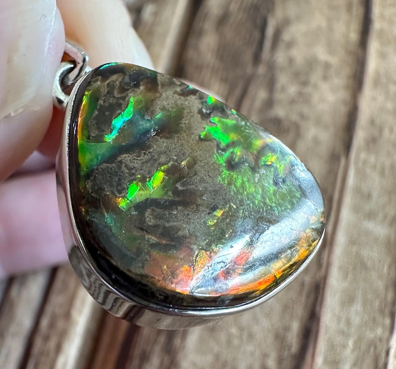 May include: A teardrop-shaped pendant with a silver setting and a dark green, iridescent gemstone. The gemstone has a mottled pattern with flashes of green, orange, and red.