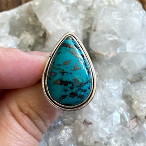 May include: A teardrop-shaped ring with a turquoise gemstone set in a silver band. The gemstone has a mottled pattern of blue and black.