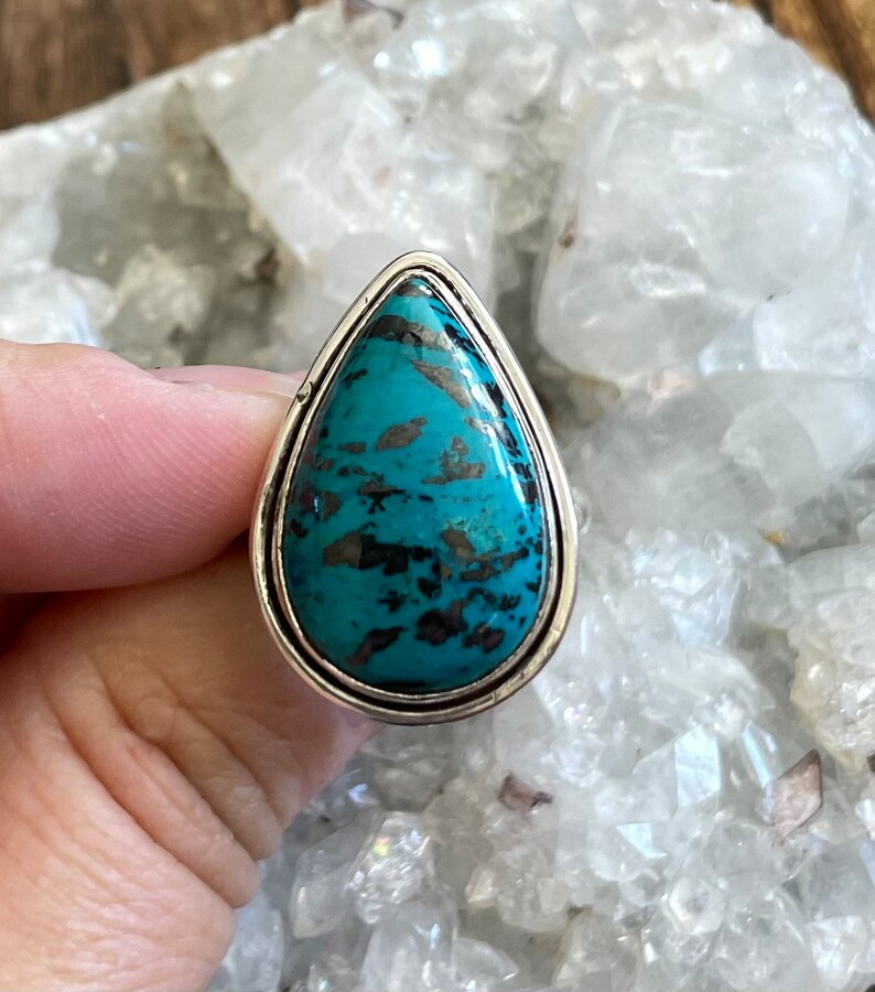 May include: A teardrop-shaped ring with a turquoise gemstone set in a silver band. The gemstone has a mottled pattern of blue and black.