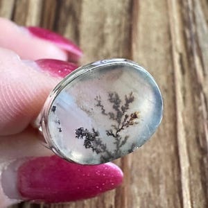 May include: A silver ring with an oval-shaped, clear stone. The stone features a dendritic inclusion resembling a tree branch in shades of black and brown. The ring is set against a blurred wooden background.