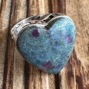 May include: A heart-shaped ring with a green and blue gemstone set in sterling silver. The gemstone has a speckled pattern with hints of pink.