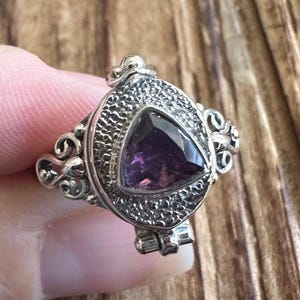 May include: A silver ring with a hinged locket featuring a triangular amethyst gemstone. The ring has an intricate, ornate design.