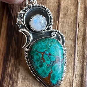 May include: A silver pendant with a green and brown gemstone and a white moonstone. The pendant is surrounded by a silver design with stars.