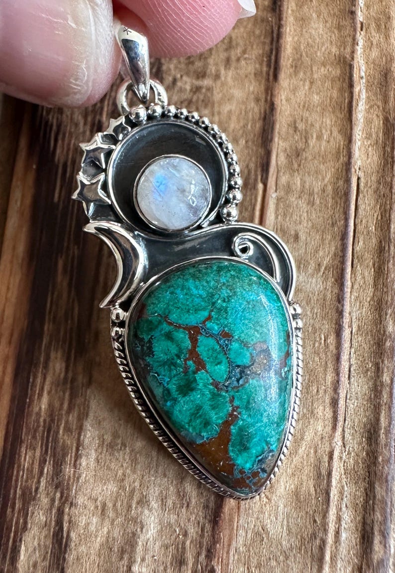 May include: A silver pendant with a green and brown gemstone and a white moonstone. The pendant is surrounded by a silver design with stars.