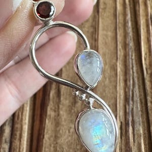 May include: A silver pendant featuring a round, dark red gemstone and two pear-shaped, iridescent moonstones. The pendant has a flowing, organic design with a small cluster of silver beads. The gemstones are set in a polished silver setting.