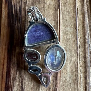 May include: A silver pendant with a variety of gemstones, including a large oval moonstone, a smaller oval garnet, and a rectangular piece of purple charoite.
