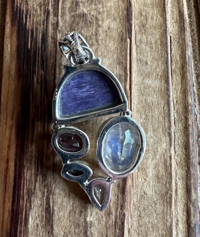 May include: A silver pendant with a variety of gemstones, including a large oval moonstone, a smaller oval garnet, and a rectangular piece of purple charoite.