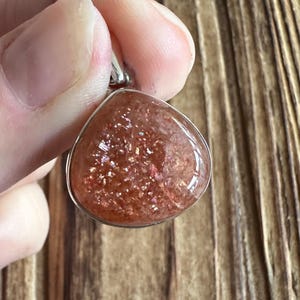 May include: A silver pendant featuring a teardrop-shaped sunstone with a reddish-orange hue and a shimmery, iridescent finish.