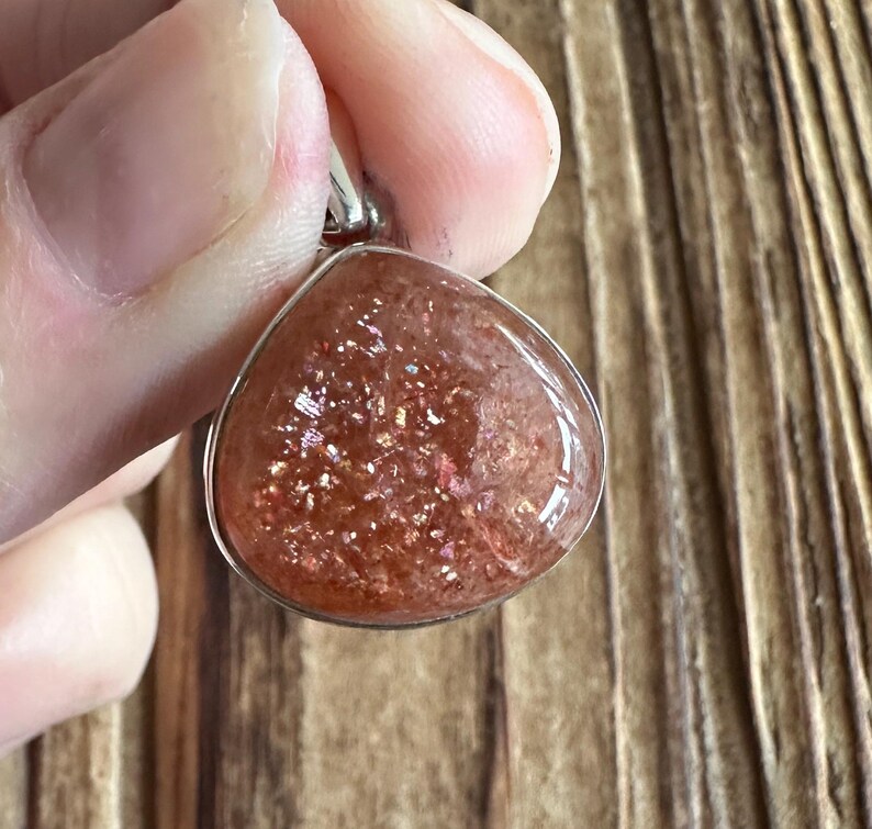 May include: A silver pendant featuring a teardrop-shaped sunstone with a reddish-orange hue and a shimmery, iridescent finish.