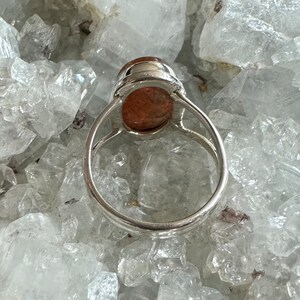 May include: A silver ring with a large, oval-shaped, reddish-brown gemstone set in the center. The ring is sitting on a bed of white crystals.