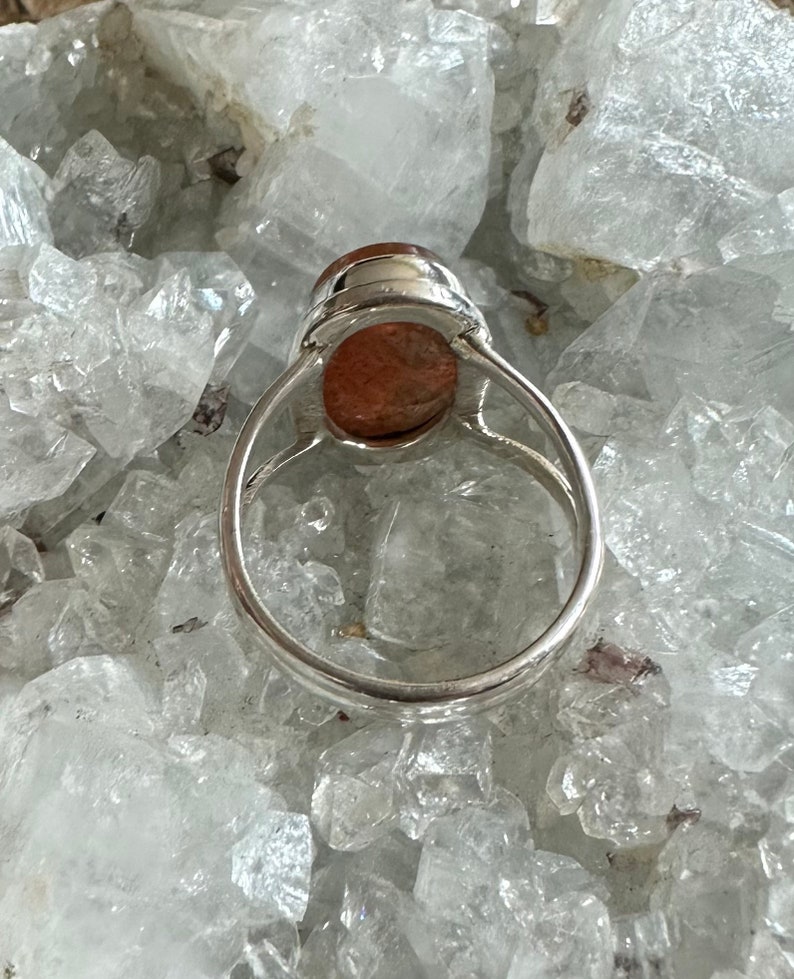 May include: A silver ring with a large, oval-shaped, reddish-brown gemstone set in the center. The ring is sitting on a bed of white crystals.