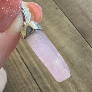 Puede incluir: Un colgante de cristal rosa p&aacute;lido con una argolla de plata. El cristal es liso y tiene una apariencia ligeramente nublada.