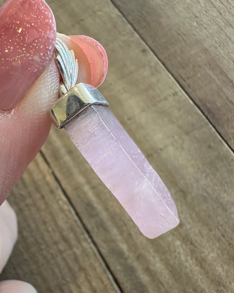 Puede incluir: Un colgante de cristal rosa p&aacute;lido con una argolla de plata. El cristal es liso y tiene una apariencia ligeramente nublada.