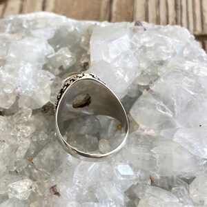 Pu&ograve; includere: Un anello d'argento con una pietra ovale di colore chiaro. L'anello ha una fascia dettagliata e decorata con motivi. L'anello &egrave; posto su uno sfondo di cristalli bianchi.