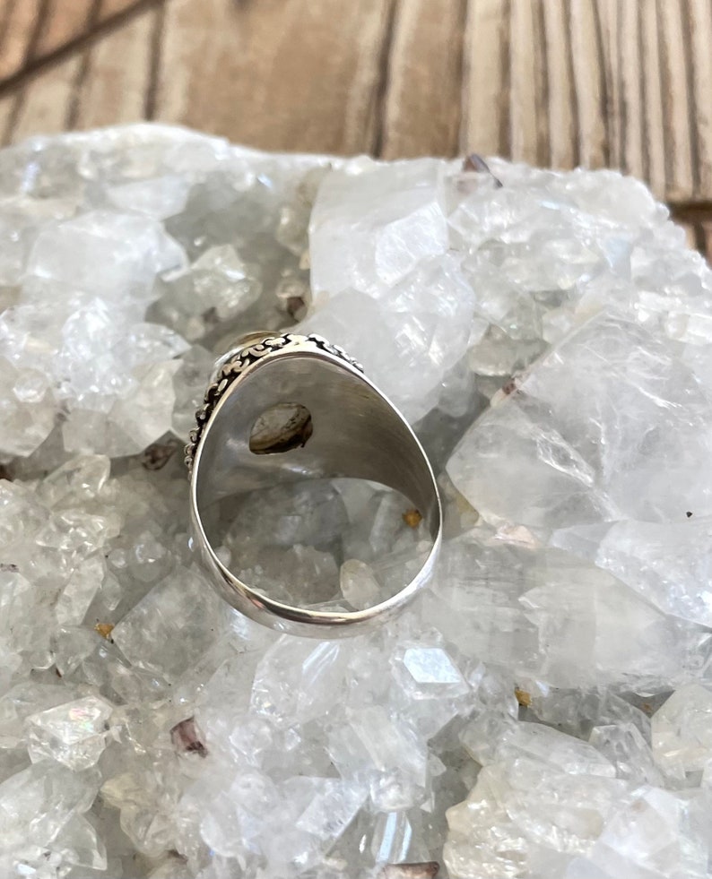 Pu&ograve; includere: Un anello d'argento con una pietra ovale di colore chiaro. L'anello ha una fascia dettagliata e decorata con motivi. L'anello &egrave; posto su uno sfondo di cristalli bianchi.