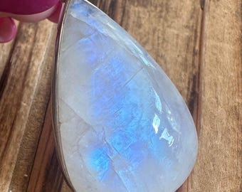 May include: A large, pear-shaped moonstone pendant with a silver-colored metal setting. The stone displays a milky white base with iridescent blue flashes. The pendant is held up by a person with pink nail polish.