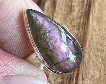 May include: A teardrop-shaped ring featuring a polished, iridescent gemstone with shades of purple, gray, and silver. The stone is set in a silver band. The ring is a piece of jewelry.