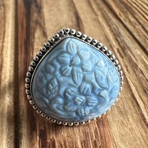 May include: A light blue gemstone ring with a floral design. The ring features a large, teardrop-shaped stone with carved leaves and flowers. The stone is set in a silver band with a beaded edge.