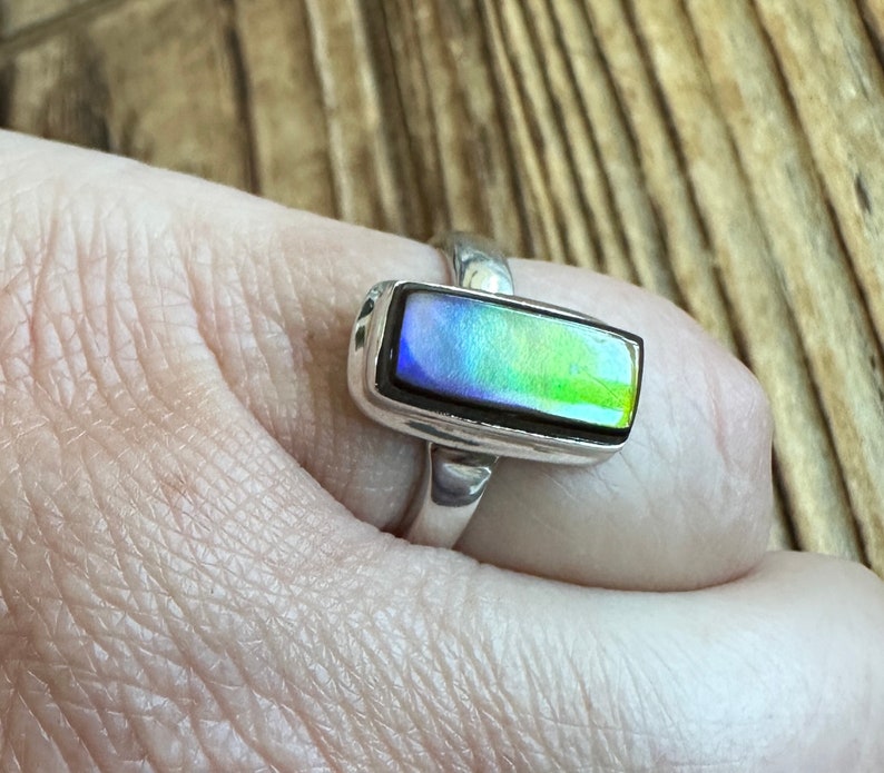 K&ouml;nnte beinhalten: Ein silberner Ring mit einem rechteckigen, schillernden Edelstein. Der Edelstein hat Blaut&ouml;ne, Gr&uuml;nt&ouml;ne und Gelbt&ouml;ne.