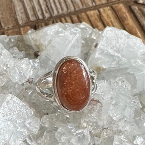 May include: A silver ring with an oval-shaped sunstone gemstone. The stone is a reddish-brown color with a shimmery, iridescent finish.