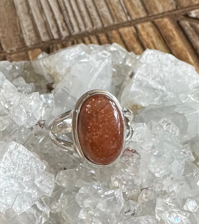 May include: A silver ring with an oval-shaped sunstone gemstone. The stone is a reddish-brown color with a shimmery, iridescent finish.