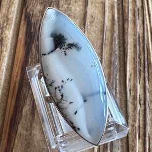May include: A large, oval-shaped ring featuring a polished, light blue gemstone with black dendritic inclusions. The stone is set in a silver band. The ring is displayed on a clear plastic stand against a wooden background.