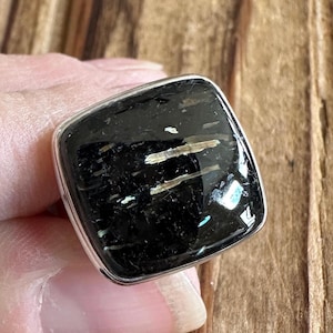 May include: A square-shaped ring with a black gemstone set in silver. The gemstone has a unique pattern of white and gold streaks.