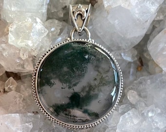 May include: A round green moss agate pendant with a silver bezel and bail. The pendant is set against a background of white crystals.