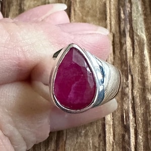 May include: A silver ring with a teardrop-shaped ruby gemstone. The ring has a textured band.
