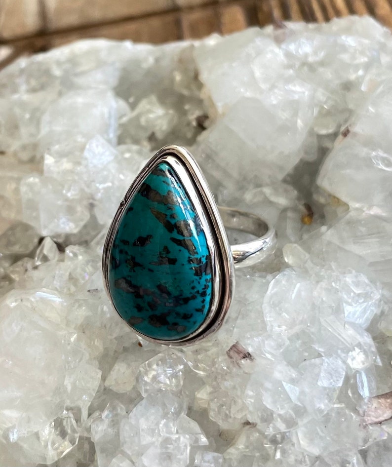 May include: A teardrop-shaped turquoise ring set in sterling silver. The turquoise is a deep blue-green color with black and white veins.