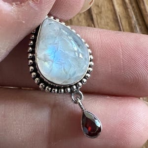 May include: A silver pendant featuring a large, pear-shaped moonstone with iridescent blue and white hues. A small, teardrop-shaped garnet dangles below. The pendant is held by a person.