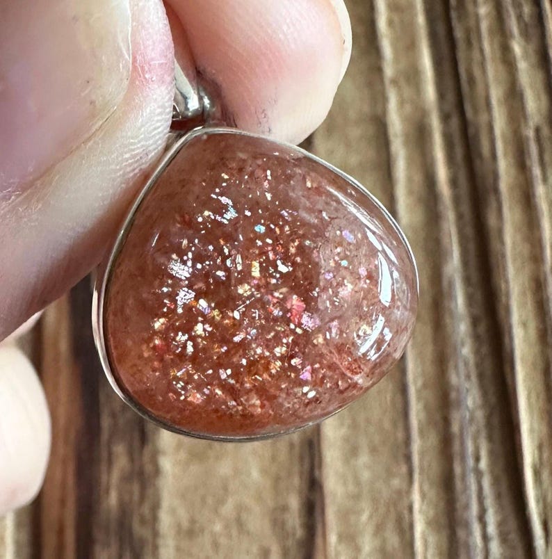 May include: A silver pendant featuring a teardrop-shaped sunstone with a shimmering, iridescent finish. The stone has a reddish-brown base with flecks of gold and silver.
