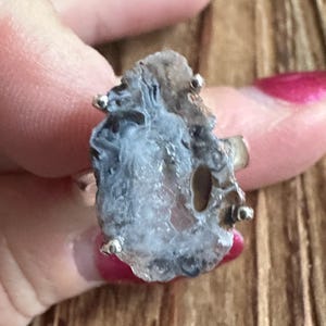 Peut inclure: Une bague unique avec une pierre brute et irrégulière, aux nuances de gris et de brun. La pierre est montée sur une monture argentée et ornée de petites perles argentées. Un bijou audacieux.