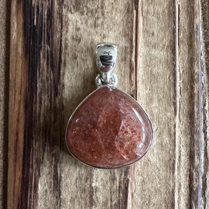 May include: A silver pendant featuring a teardrop-shaped sunstone with a reddish-brown hue and a shimmery, iridescent finish.