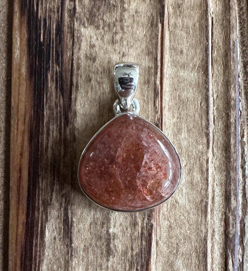 May include: A silver pendant featuring a teardrop-shaped sunstone with a reddish-brown hue and a shimmery, iridescent finish.